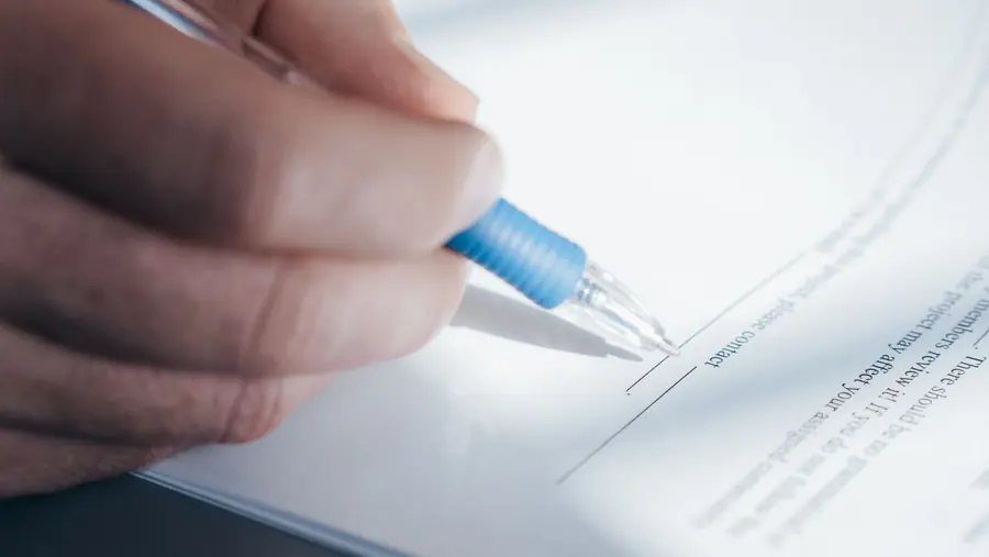A person signing a document with a pen on a desk.